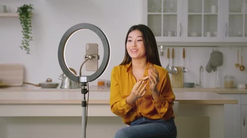 Joven Blogger de belleza asiática grabando Vídeo sobre el nuevo colorete sentado en la cocina con teléfono inteligente