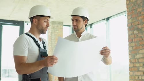 Dois jovens engenheiros em capacetes de segurança com um plano de projeto em pé no canteiro de obras