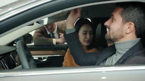 Man and Woman with Child Sitting Inside Car Choosing Auto in Showroom Talking To Manager