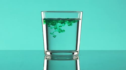 Drops of Green Ink Dripping Into Clear Water in Glass Standing on Turquoise Background in Slowmotion