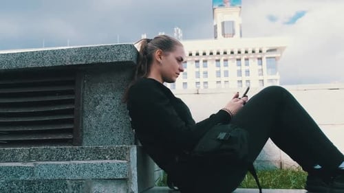 Young Woman Uses Smartphone While Sitting on the Parapet in the Street Park