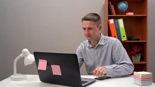 Young Guy Office Worker Manager is Typing Text in the Phone and in the Computer