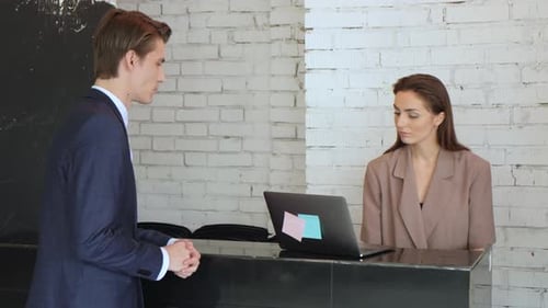 Man in a strict suit approaches a reception in hotel and talks to the girl