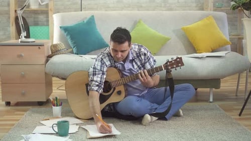 Man Playing Guitar and Writing Song at Home