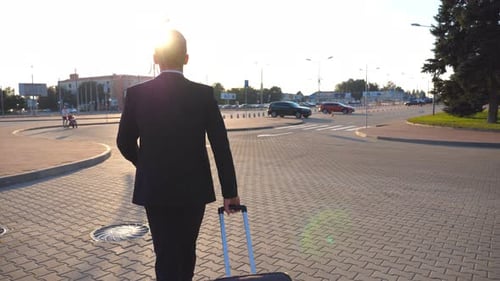 Rear View of Unrecognizable Young Businessman Walking to Airport Terminal and Pulling Suitcase on