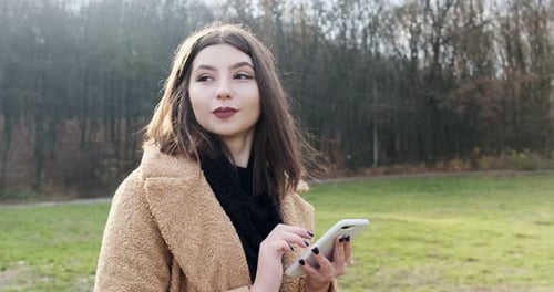 Portrait of Young Attractive Woman Smiling and Looking at Smartphone on the Green Lawn at Autumn