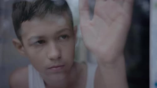 Sad Boy Stands at Window with Hand on Glass