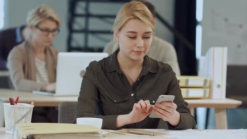 Cheerful Young Woman Using Her Smartphone with Smile While Sitting at Her Working Place