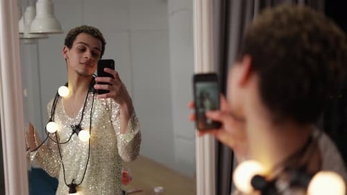 Um homem em um vestido posando na frente do espelho com guirlanda no pescoço e copo de vinho usando telefone