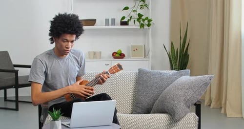 Young Adult Plays Ukulele While Using Laptop