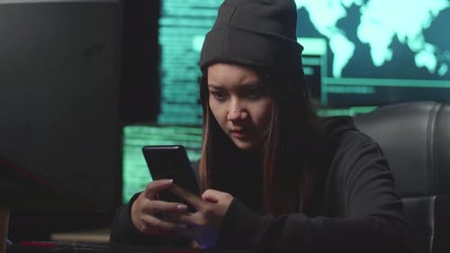 Hacker asiática que usa un teléfono móvil durante el hackeo de una computadora, Código en varias pantallas de computadora