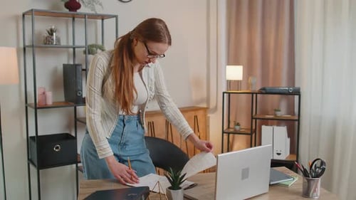 Young Woman Girl Standing By Desk in Home Office Working at Paper Documents Analyzes Checks Data