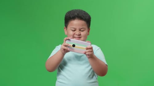 Excited Asian Little Boy Playing Video Game With Mobile Phone In Green Screen Studio