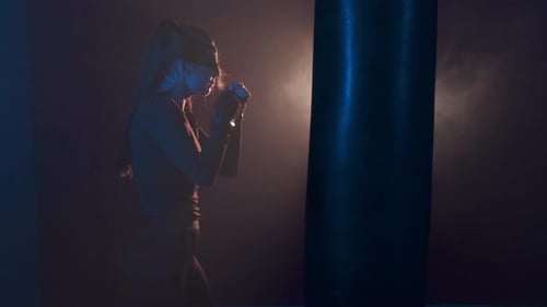 Beautiful strong woman kickboxer alone is engaged in a dark sports hall, beats a punching bag.