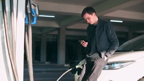 Young Adult Using Phone While Charging Electric Car