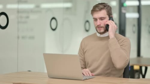 Young Adult Using Laptop, Talking on Phone at Work