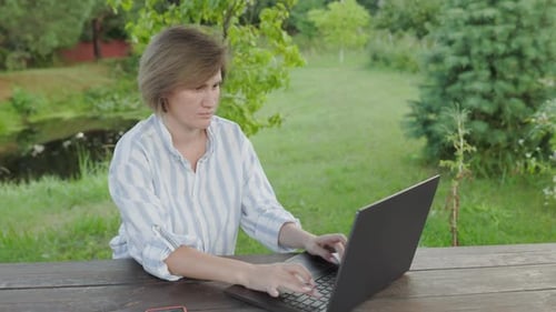 woman is sitting with a laptop and typing on it in the garden of a country house.