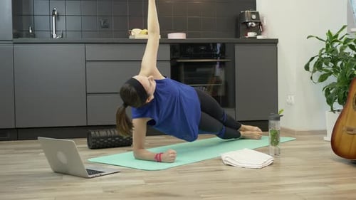 Woman in athlete sports clothes watching online fitness clases, doing side plank on mat in kitchen