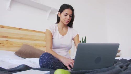 Fröhliche junge Frau, die morgens zu Hause mit Laptop im Bett arbeitet