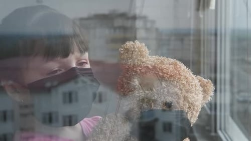 Little Girl With Mask Looking Out Window Holding Teddy