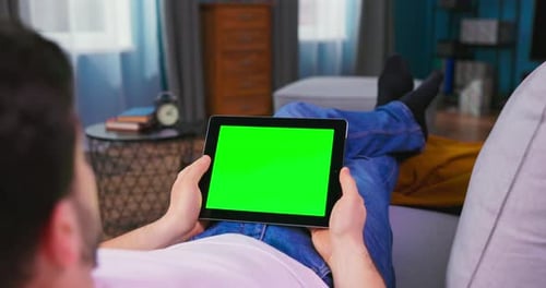 Young Man at Home Laying on a Couch Using with Green Mockup Screen Tablet Computer