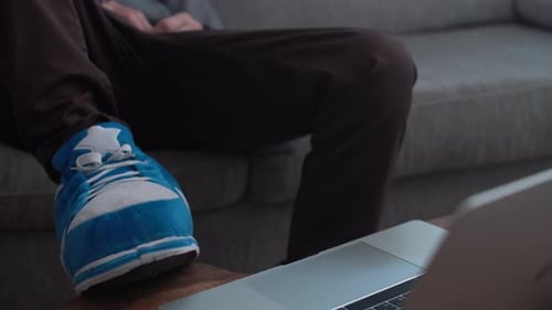 Homem com chinelos senta-se confortavelmente no sofá e depois digita no laptop