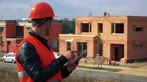 Ein junger Bauarbeiter arbeitet auf einem Tablet, im Hintergrund ein im Bau befindliches Haus
