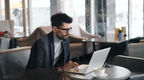 Männlicher Freelancer arbeitet im Café mit Laptop und schaut auf den Bildschirm und berührt die Tastatur
