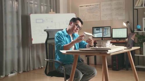 Asian Man Engineer With A Laptop Thinking While Holding Paper Model Of House At The Office