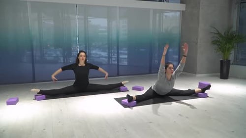 Two Athletic Women Stretching Legs, Leans Forward Sitting on a Splits on Mat in the Gym
