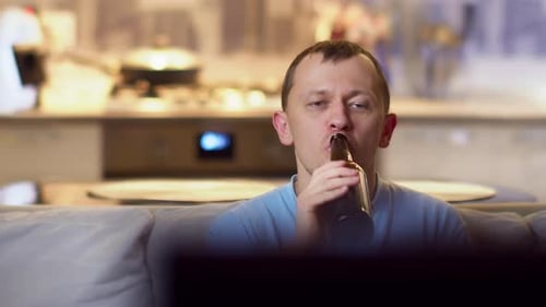 Young man drinking beer from a bottle, sitting at home on the sofa and watching TV, camera movement
