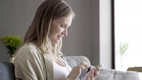 Woman Using Smartphone on Sofa with Earbuds