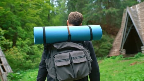 A Young Man Traveling with a Backpack Walks Against the Backdrop of a Forest Hut