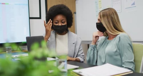 Two diverse businesswomen wearing face masks working together using laptop at desk in office