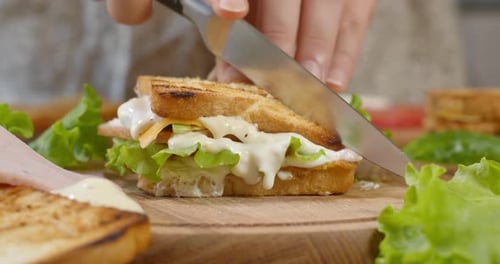Female Chef Cuts Club Sandwich in a Half By Sharp Knife on a Wooden Board at the Kitchen