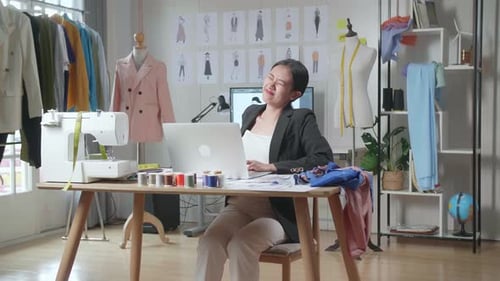Diseñadora en traje de negocios que trabaja en una computadora portátil y tiene dolor de espalda mientras diseña ropa