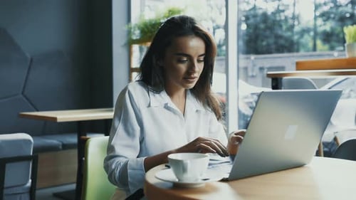 Pretty Woman in Formal Clothes Typing on Computer in Modern Cafe with Cup of Coffee