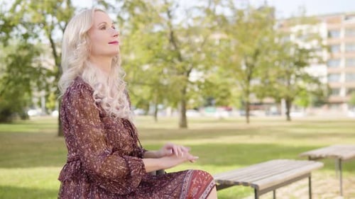 A Middleaged Caucasian Woman Looks Around with a Smile As She Sits on a Bench in a Park
