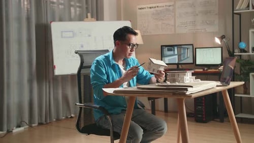 Asian Man Engineer With A Laptop Thinking While Holding Paper Model Of House At The Office