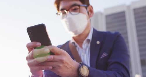 Hombre asiático con máscara facial con teléfono inteligente mientras está sentado en las escaleras del parque Corporativa