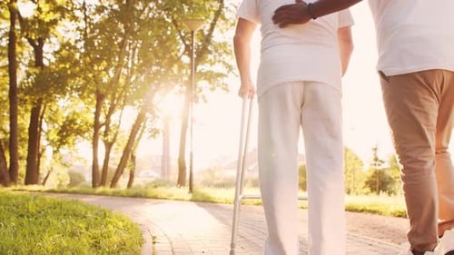 African-American caregiver is teaching disabled old man to walk with walker. Nurse and patient.