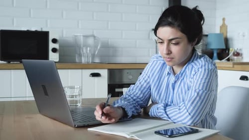 Indian Girl makes Notes in a Notebook and Works on a Computer sitting In the Kitchen at Home