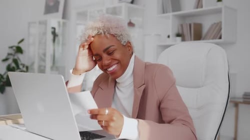 Young African American Woman Holds Piece of Paper Sits at Table in Office