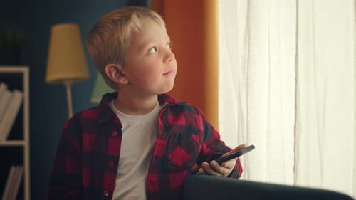 Blond Boy Gazing out Bright Window with Phone