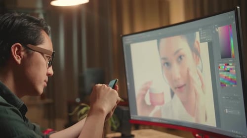 Asian Male Digital Editor Using Mobile Phone While Works In Photo Editing Software On Computer