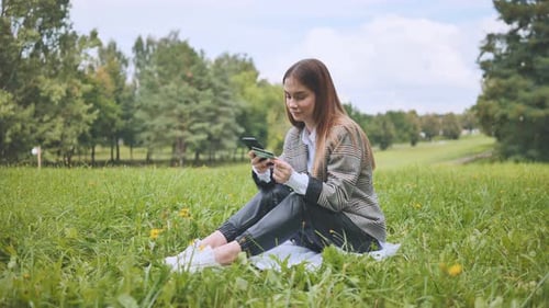 A Young Girl Makes a Purchase with Her Phone and Bank Card in the Park Sitting on the Grass