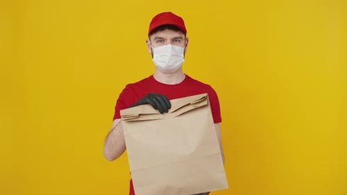 Junger Zusteller in roter Uniform mit medizinischer Schutzmaske bietet Öko-Papierpaket an