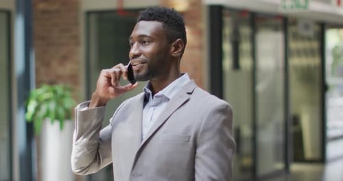 Smiling african american businessman standing in corridor talking on smartphone in modern office