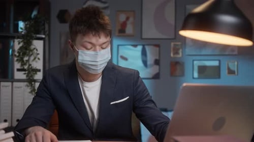 Masked Young Adult Working on Laptop in Office