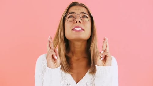 Young Lady in Glasses is Crossing Her Fingers Making a Wish Saying Please and Smiling Posing on Pink